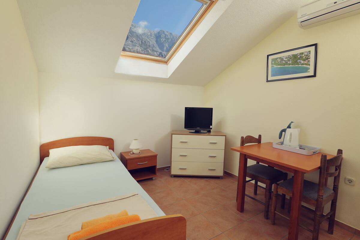 Ein kleiner, gut beleuchteter Raum mit einem Bett, einem Schreibtisch mit Stuhl, einem Fernseher auf einer Kommode und einem Fenster mit Blick auf die Berge.