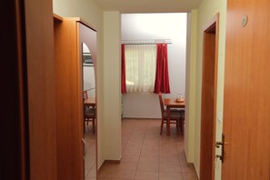 A narrow hallway with wooden doors, a dining area with a table and chairs, and a window with red curtains. - Apartments Vila Adrijana and Fitness Studio Wolf (Baska Voda)