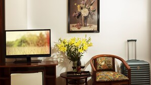 Ein Zimmer mit Fernseher, einer Vase mit gelben Blumen, einem gerahmten floralen Gemälde, einem Sessel und einem Koffer.