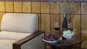 Eine Wand mit Holzvertäfelung, ein beige gepolsterter Sessel, ein Glastisch mit einer Weinflasche, einem Glas Rotwein, einer Platte mit Trauben und einer Vase mit weißen Blumen.