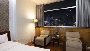 Ein Hotelzimmer mit einem Bett, zwei Sesseln, einem kleinen Tisch mit einer Vase und Weingläsern sowie einem Blick auf die Stadt durch ein großes Fenster.