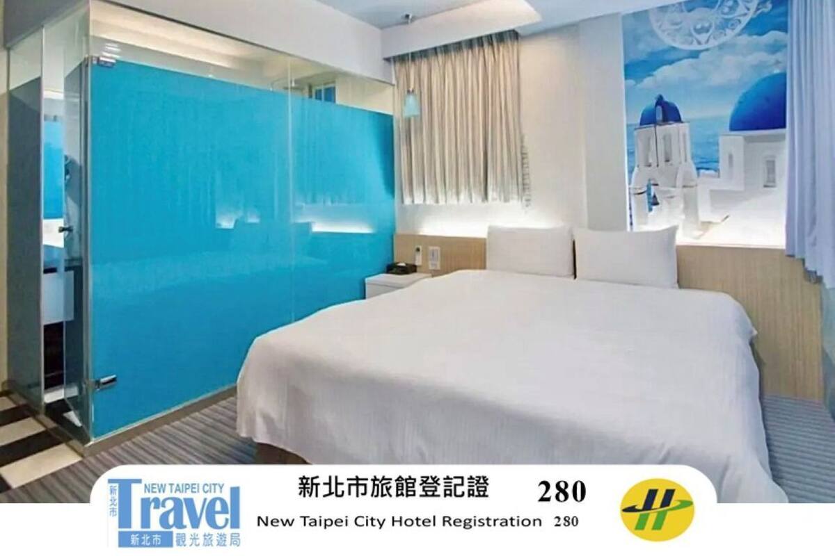 A modern hotel room with a large bed, a glass partition, and a mural of a blue sky and white buildings.