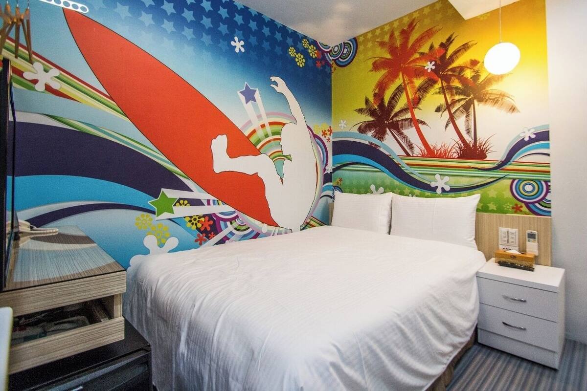 A hotel room with a bed, a nightstand, and a wall mural featuring a surfer and palm trees.