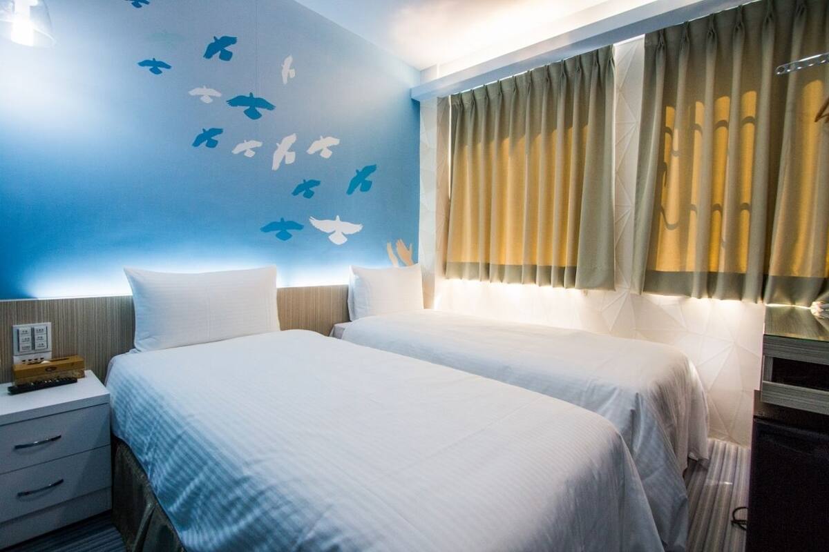 A hotel room with two beds, a wall with bird decals, a window with curtains, and a bedside table with a lamp.