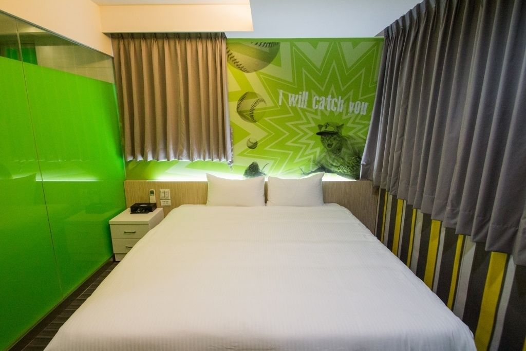 A hotel room with a bed, a nightstand, a lamp, and a green wall with a baseball-themed mural.