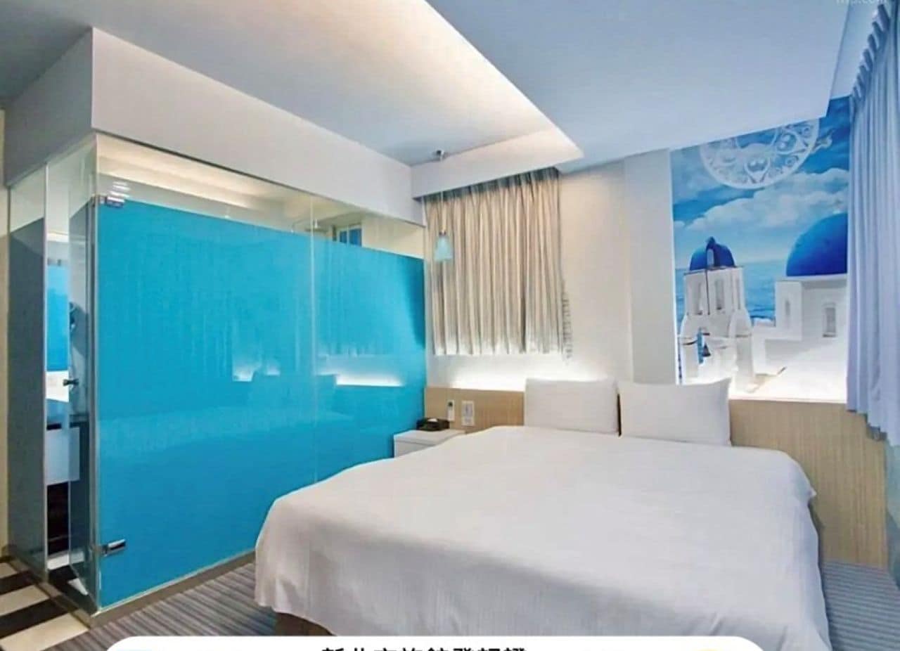 Ein modernes Hotelzimmer mit einem großen Bett, einer Glastrennwand und einem Wandbild mit blauem Himmel und weißen Gebäuden.