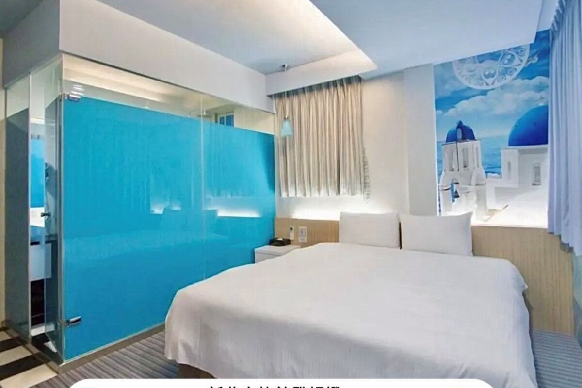 A modern hotel room with a large bed, a glass partition, and a mural of a blue sky and white buildings.