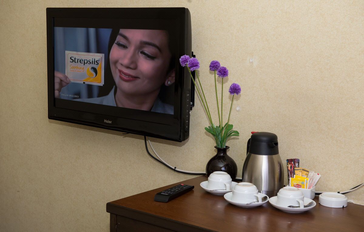 Un televisor muestra a una persona sosteniendo una caja de StrepSils, una cafetera, un florero con flores moradas y una mesa con tazas y un control remoto.