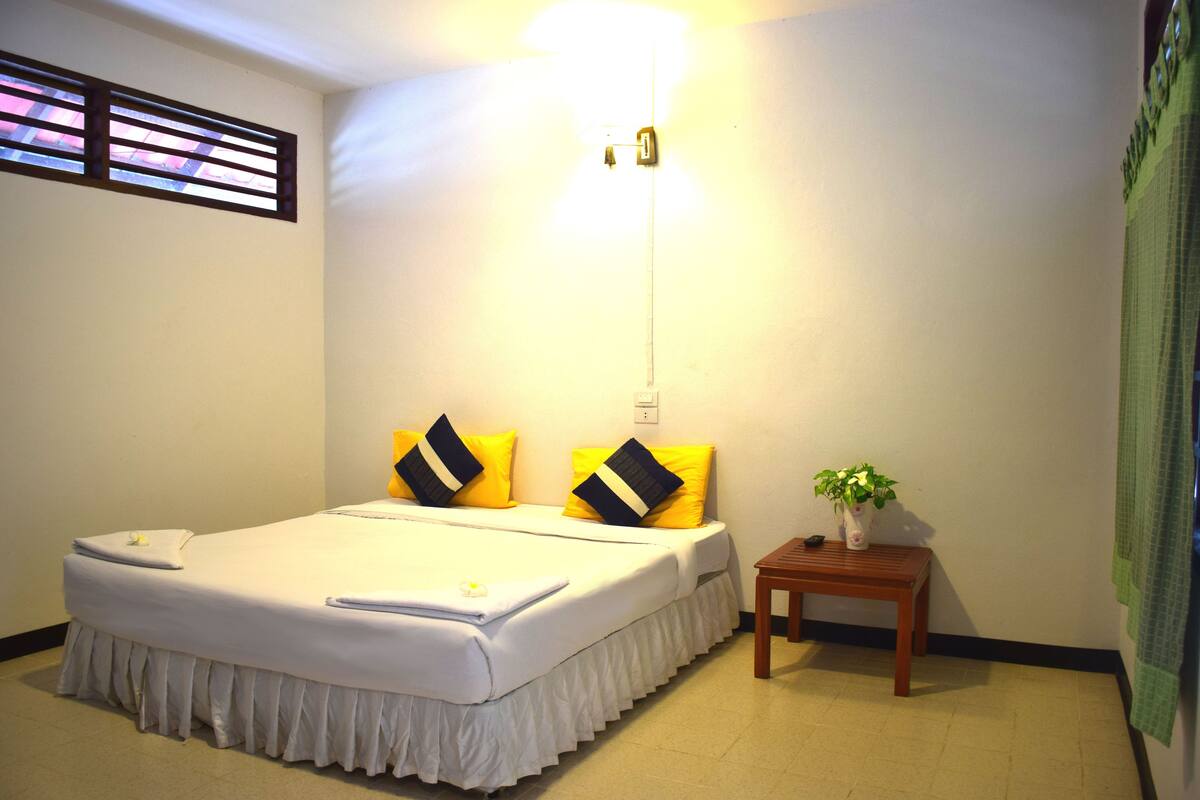 A hotel room with a bed, a bedside table with a plant, and a window with blinds.