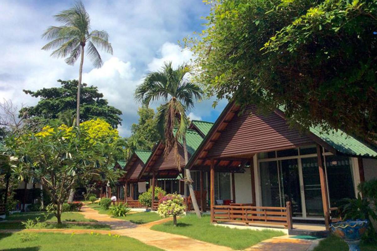 A row of wooden bungalows with green roofs, surrounded by lush greenery and palm trees.