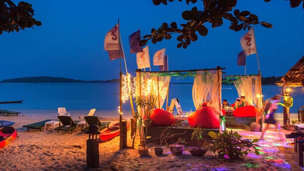 Un petit chalet au bord de la plage, doté d’un auvent et entouré de plantes en pot et de poufs rouges.