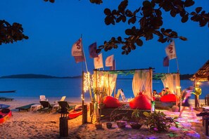 Una cabaña junto a la playa con un toldo, rodeada de macetas con plantas y pufs rojos.