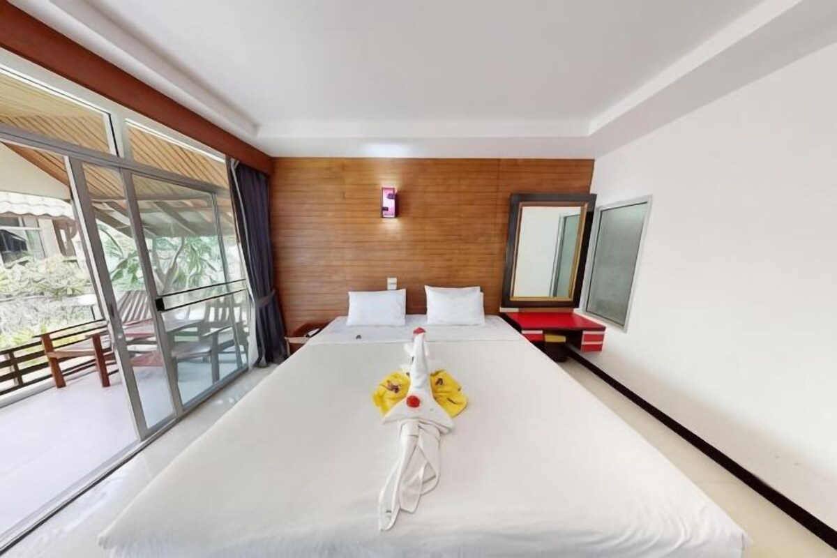 Een hotelkamer met een groot bed, een schuifpui naar buiten, een houten wand en een klein tafeltje met een rood voorwerp erop.
