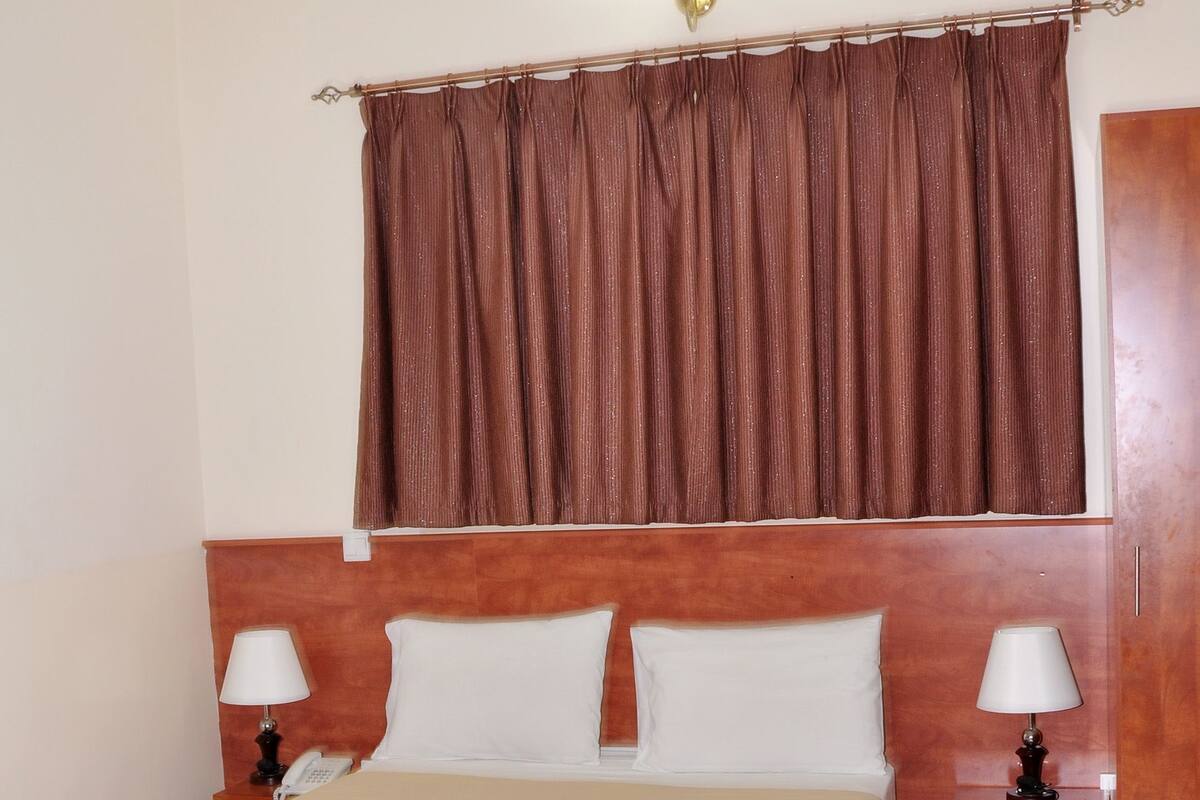 A hotel room with a large bed, two bedside tables with lamps, a wooden headboard, a brown curtain, and a small rug.