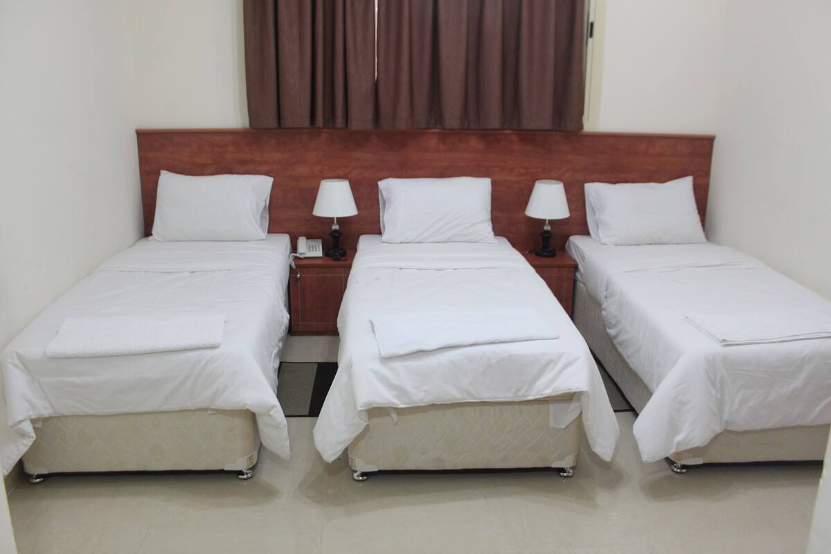 A hotel room with three single beds, each with a white bedspread and pillow, a wooden headboard, and bedside lamps.