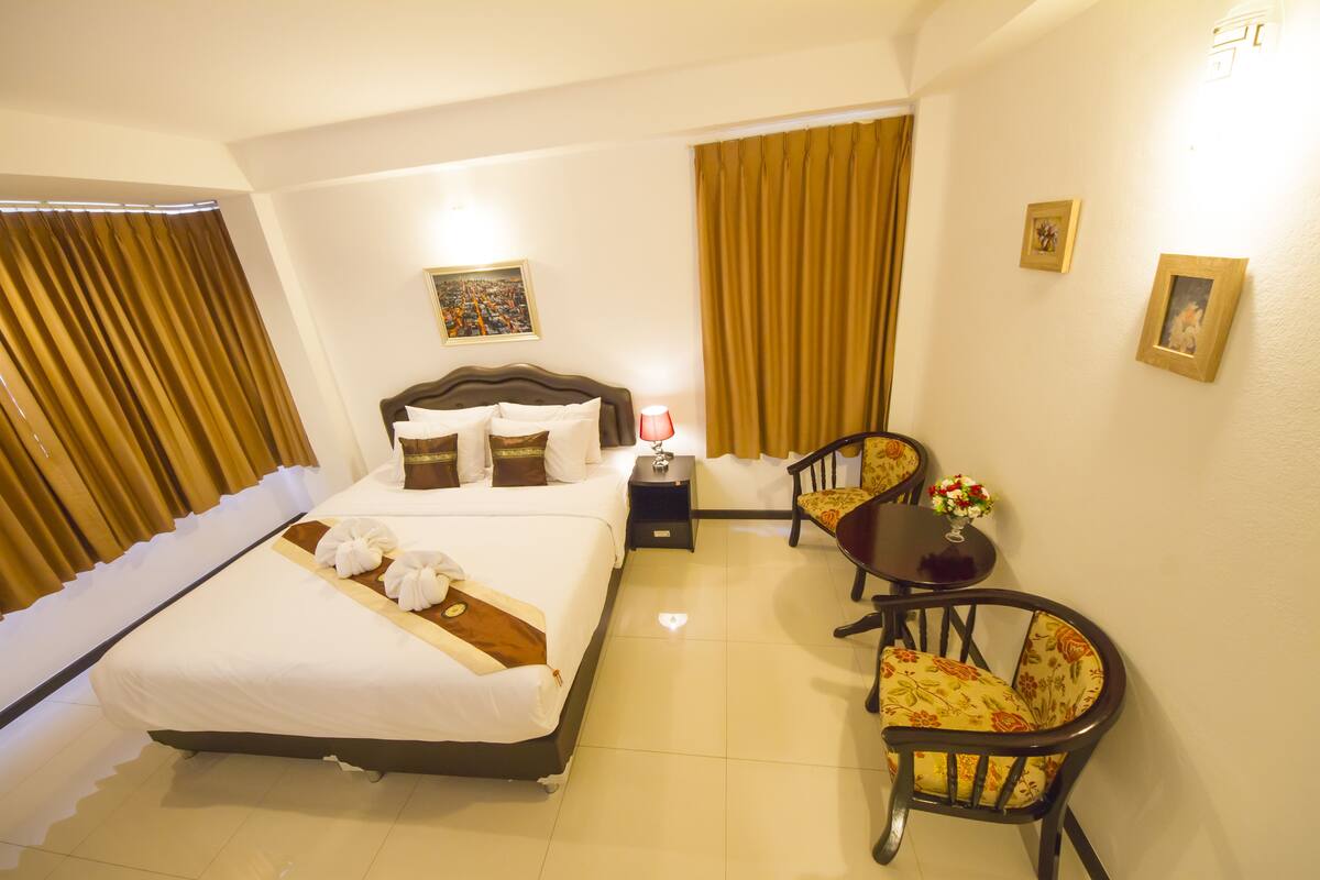 Une chambre d’hôtel avec un lit, une table de chevet, une lampe, deux chaises, une petite table avec des fleurs et des rideaux.
