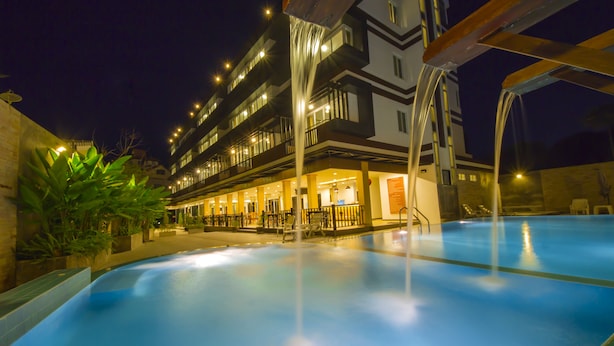 A swimming pool with a water feature, surrounded by a building with multiple balconies and lights. - TH Beach Hotel (Hua Hin)