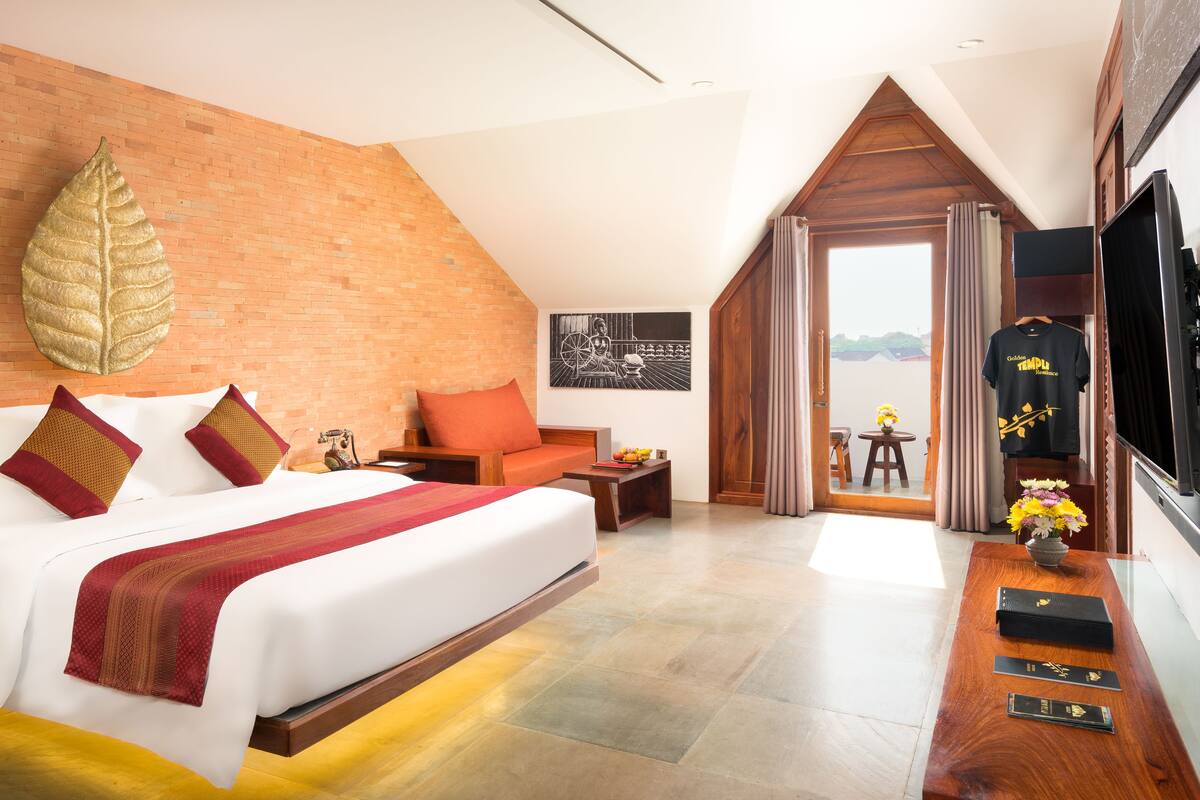 A hotel room with a large bed, a wooden bench, a small table, and a view of the outside.