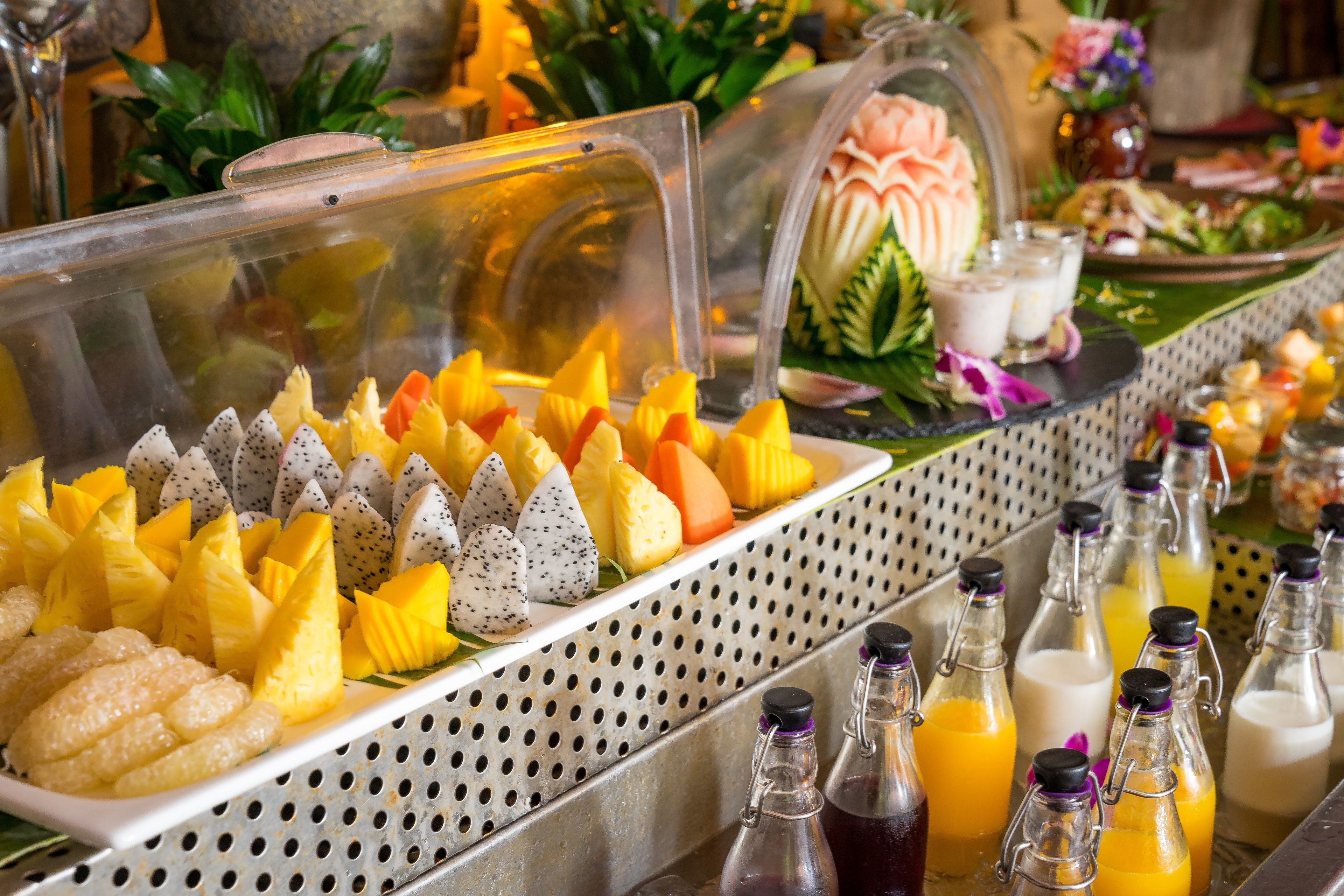 Buffet com diversas frutas, incluindo abacaxi, pitaya e manga, e várias garrafas de bebidas.