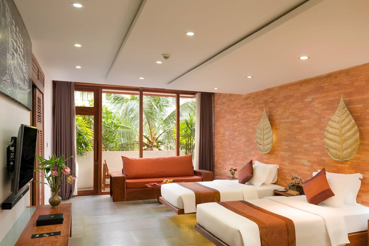 A modern hotel room with two beds, a television, a vase with flowers, and a view of greenery outside.