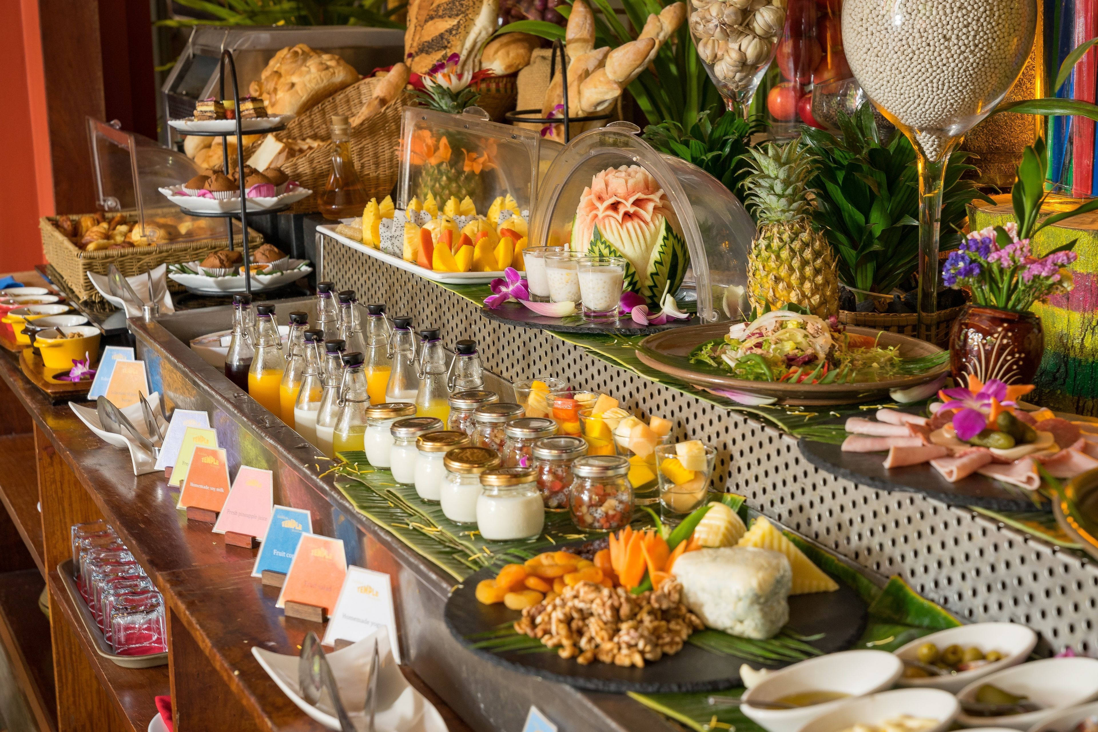 Un bufé con diversos alimentos y bebidas, que incluye frutas, pasteles y una decoración central.