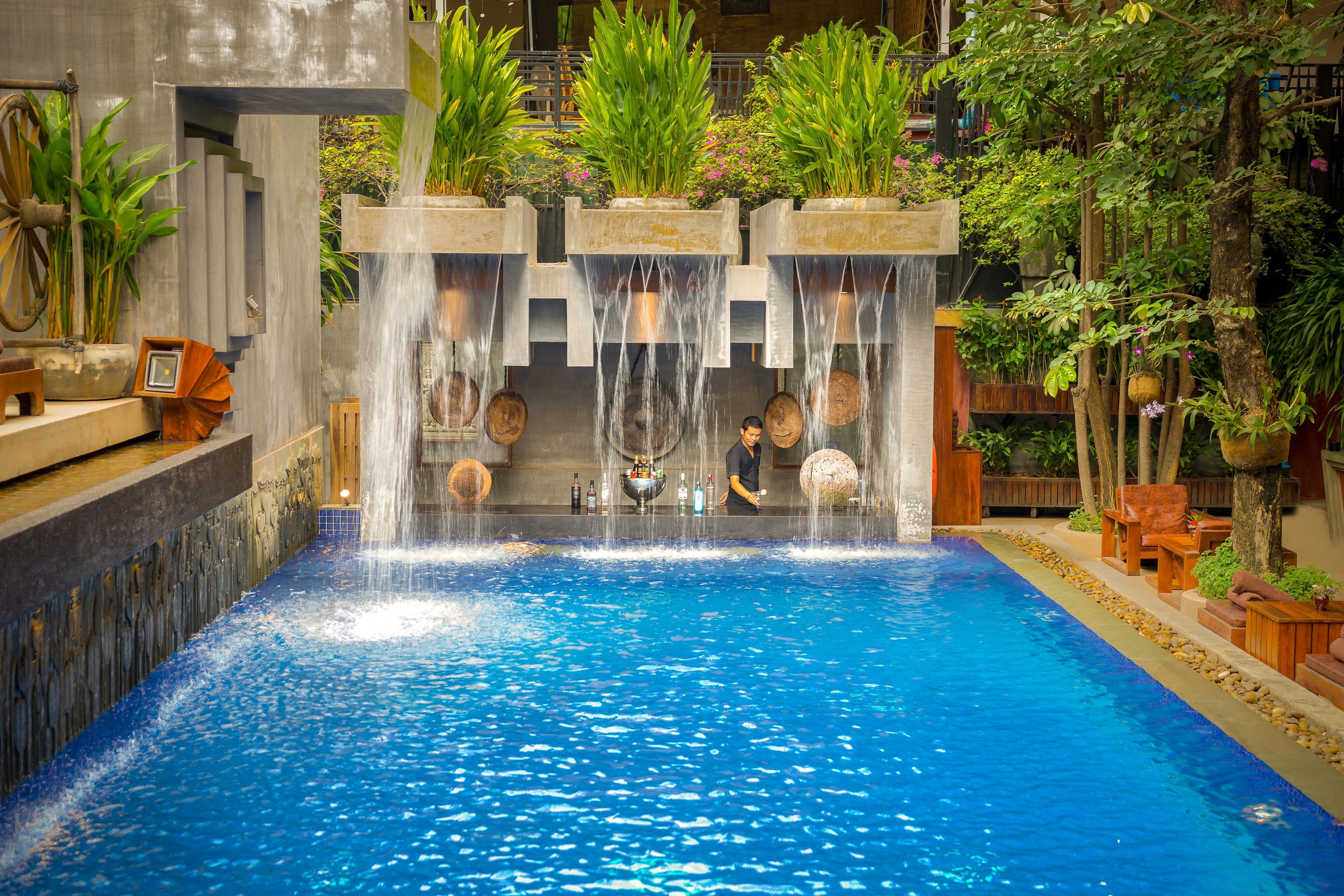 An indoor swimming pool with multiple water features, including a waterfall and fountains, surrounded by lush greenery and wooden elements.