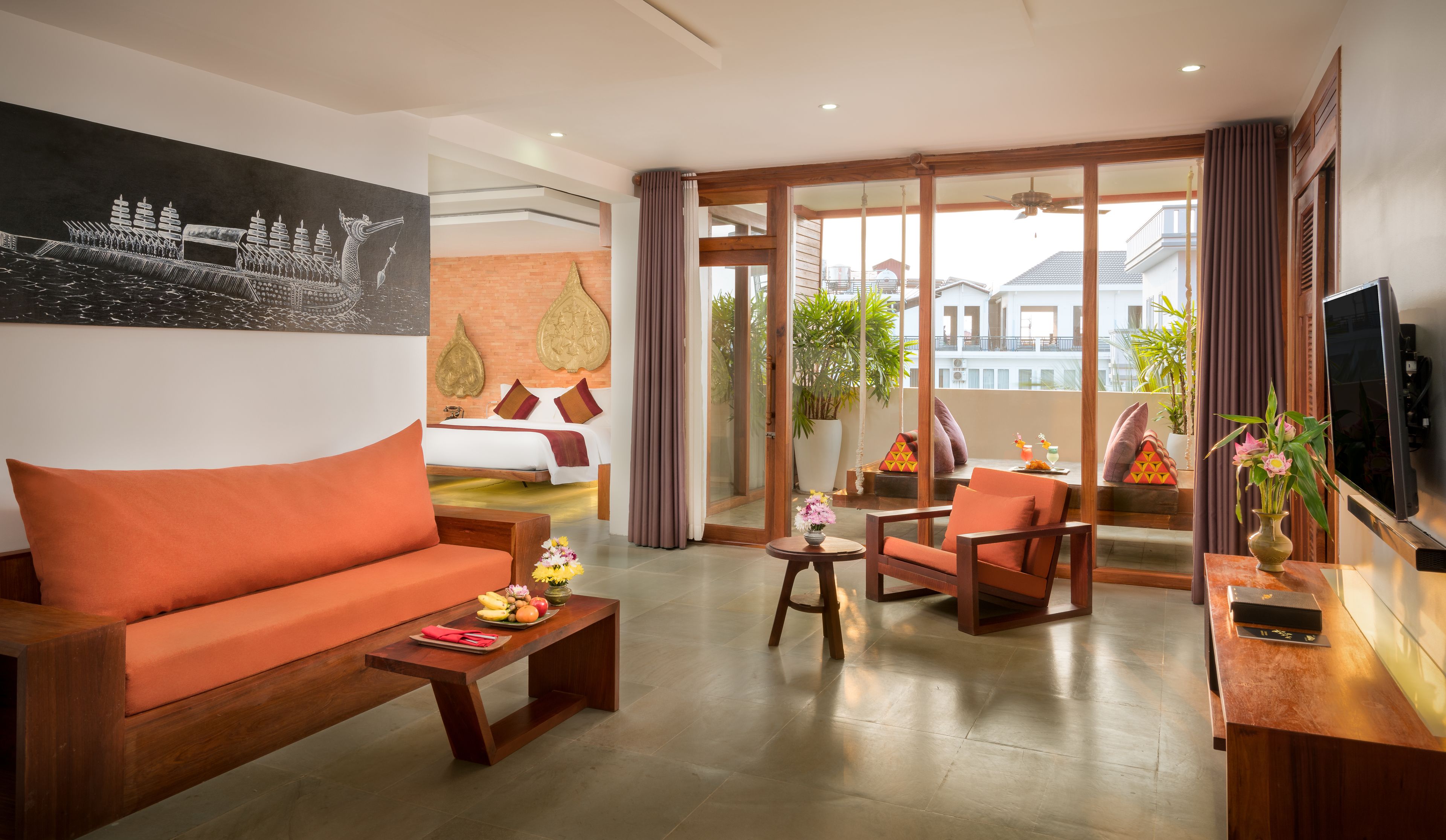 A modern hotel room with a large orange sofa, a wooden coffee table, a TV, and a balcony with a view of the city.