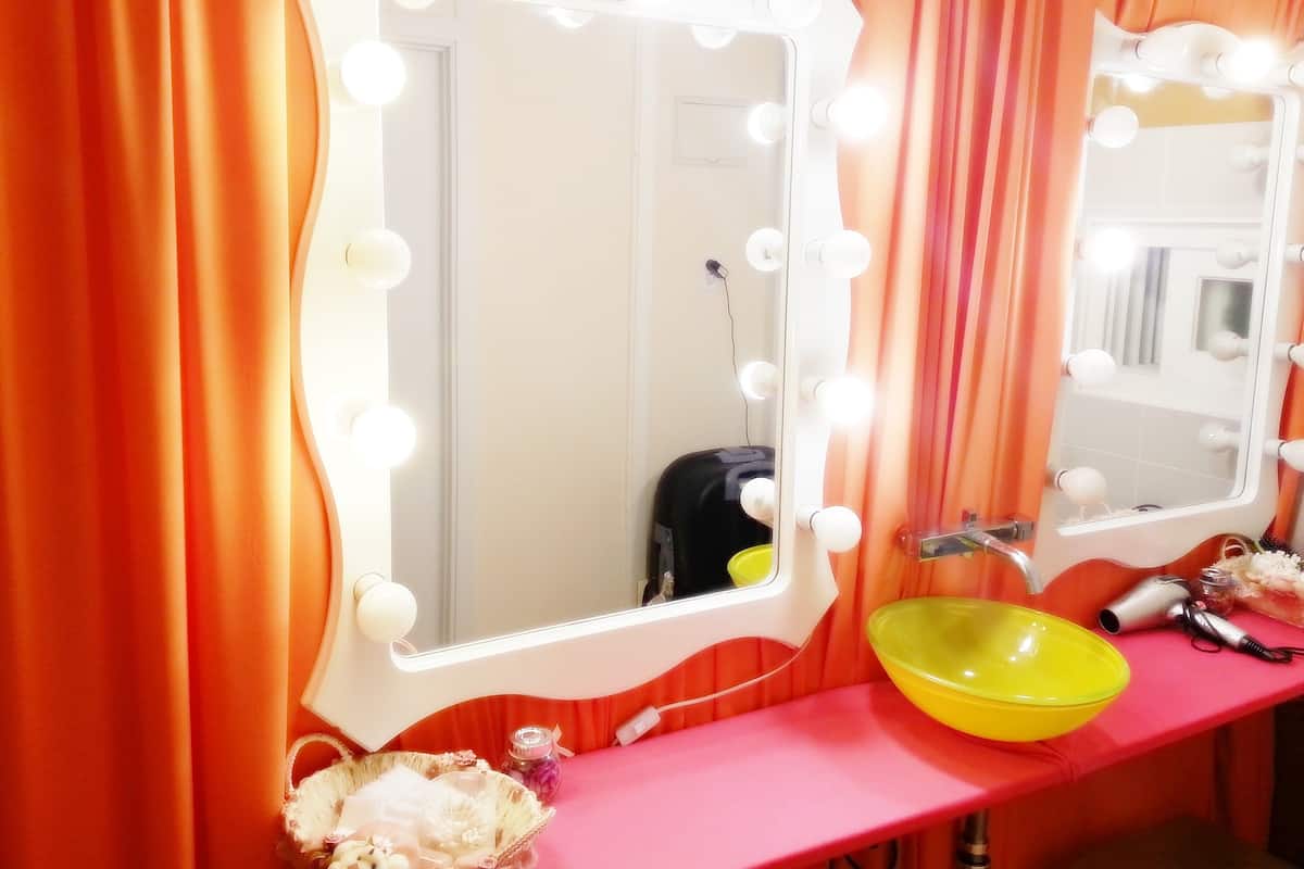 A vanity area with two mirrors surrounded by lights, a pink countertop, and a yellow bowl.