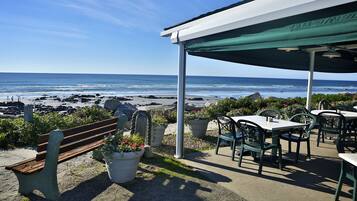 Eine Terrasse direkt am Strand mit einer Bank, Tischen und Stühlen unter einem Sonnensegel, mit Blick auf den Ozean.