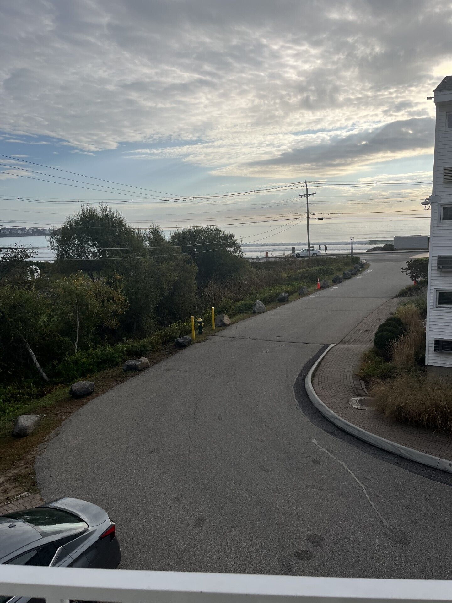 En väg som svänger åt höger, med en vit byggnad på höger sida, en vattenyta i bakgrunden och en bil parkerad på vänster sida.