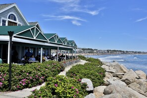 A seaside restaurant with outdoor seating, a view of the ocean, and a rocky shoreline.