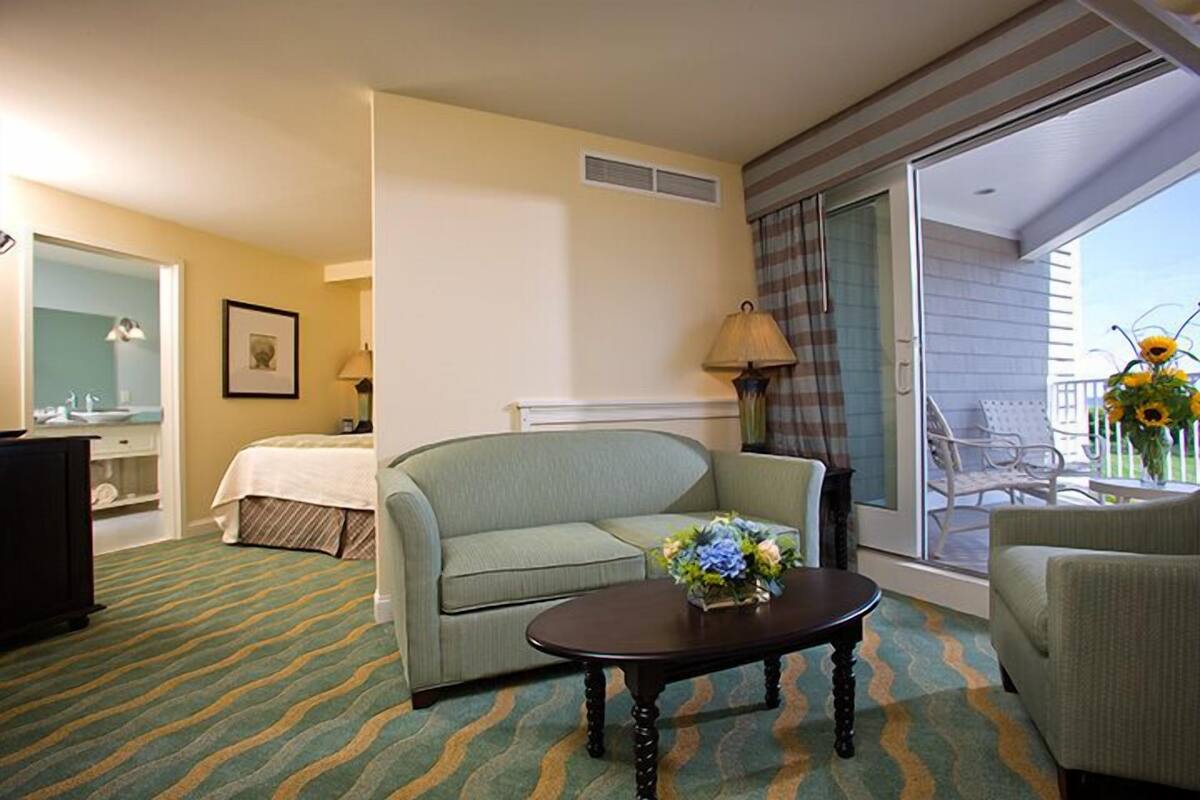 A hotel room with a bed, a sofa, a coffee table with flowers, a chair, and a balcony with a view.