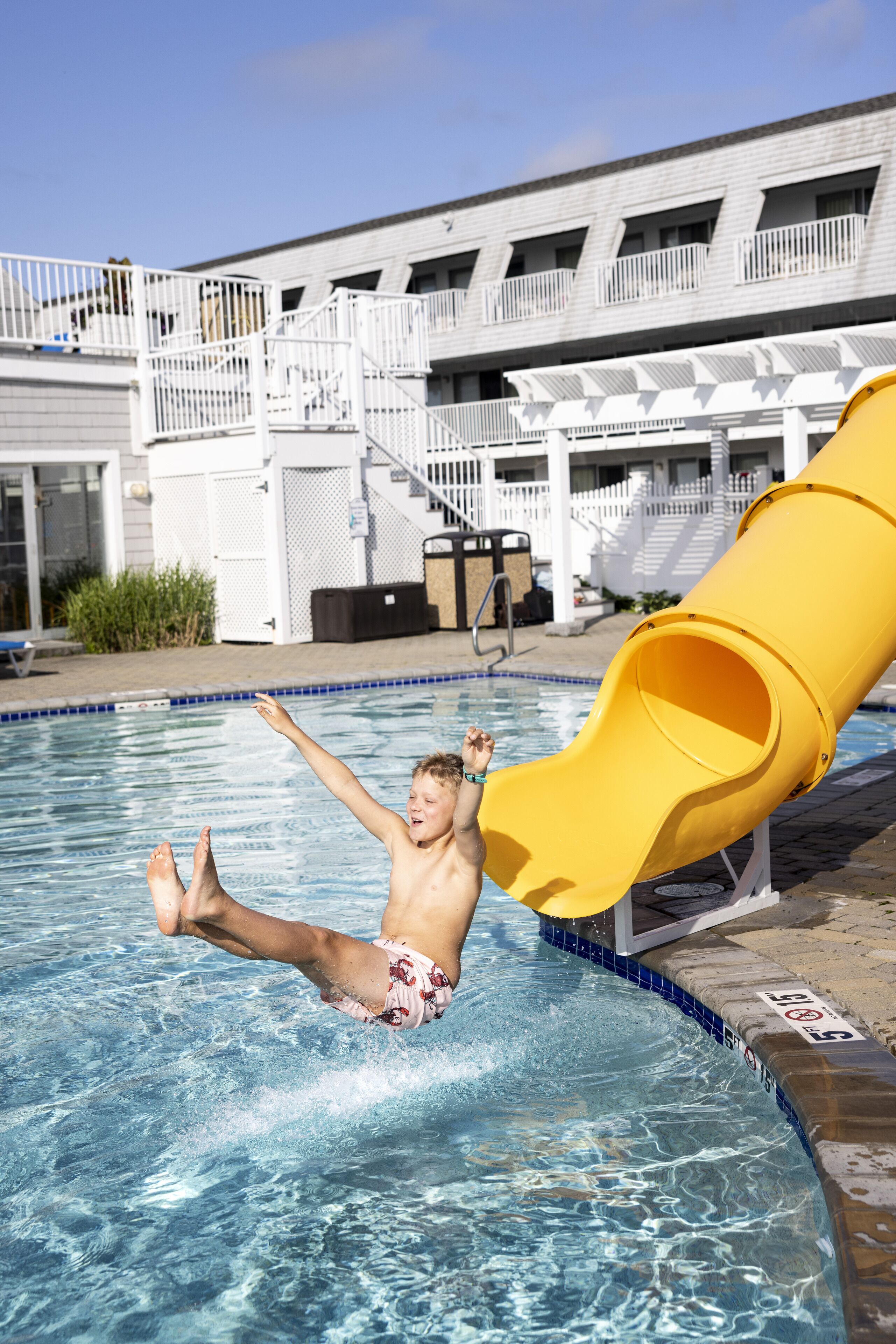 Una piscina con un tobogán amarillo, una cerca blanca y un edificio con varios balcones al fondo.