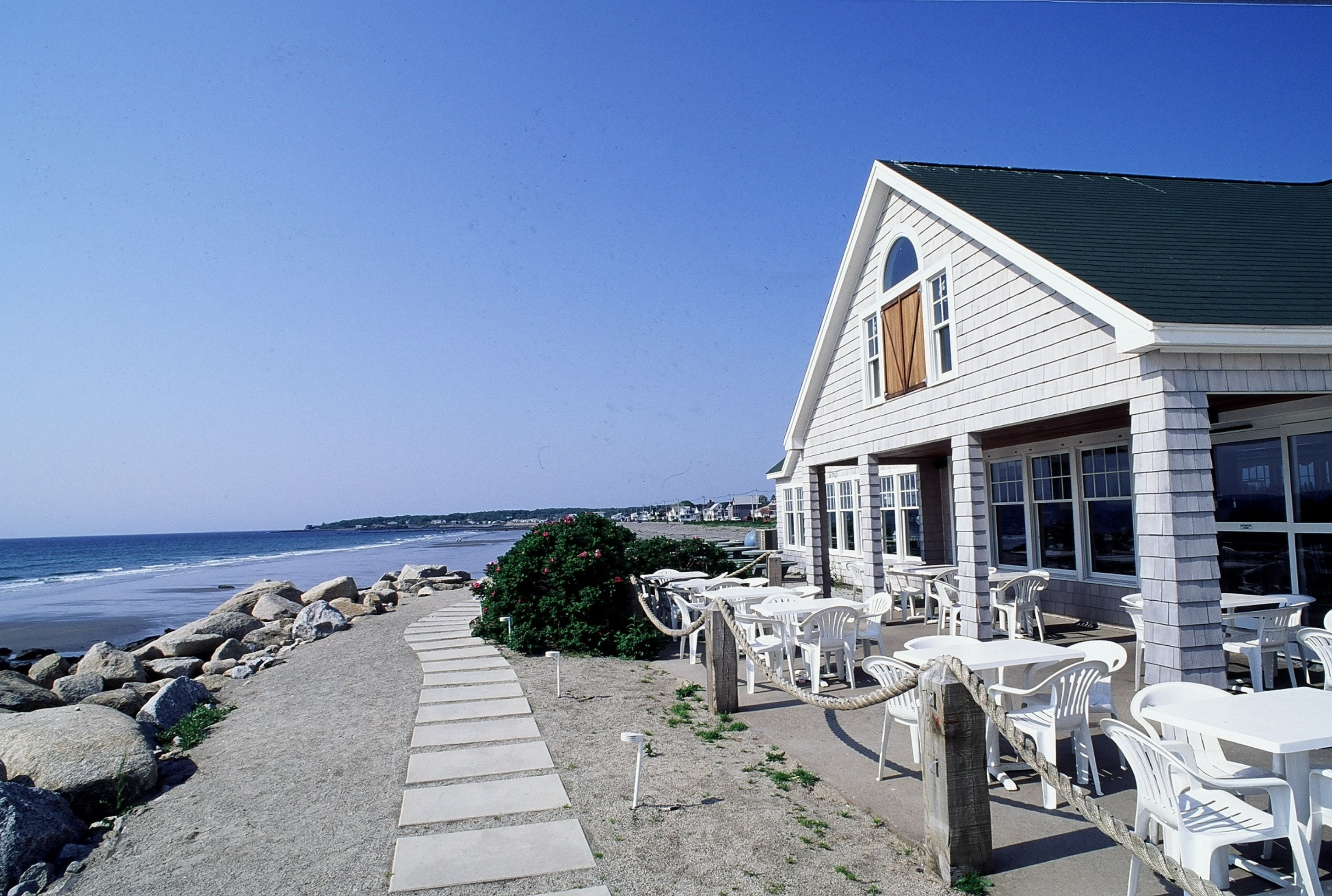 A seaside building with outdoor seating and a stone pathway leading to the beach.