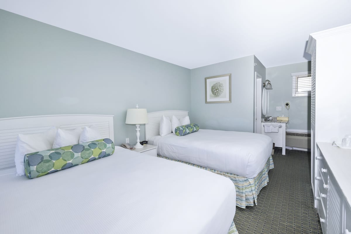 A hotel room with two beds, a bedside table with a lamp, a small bathroom, and a dresser.
