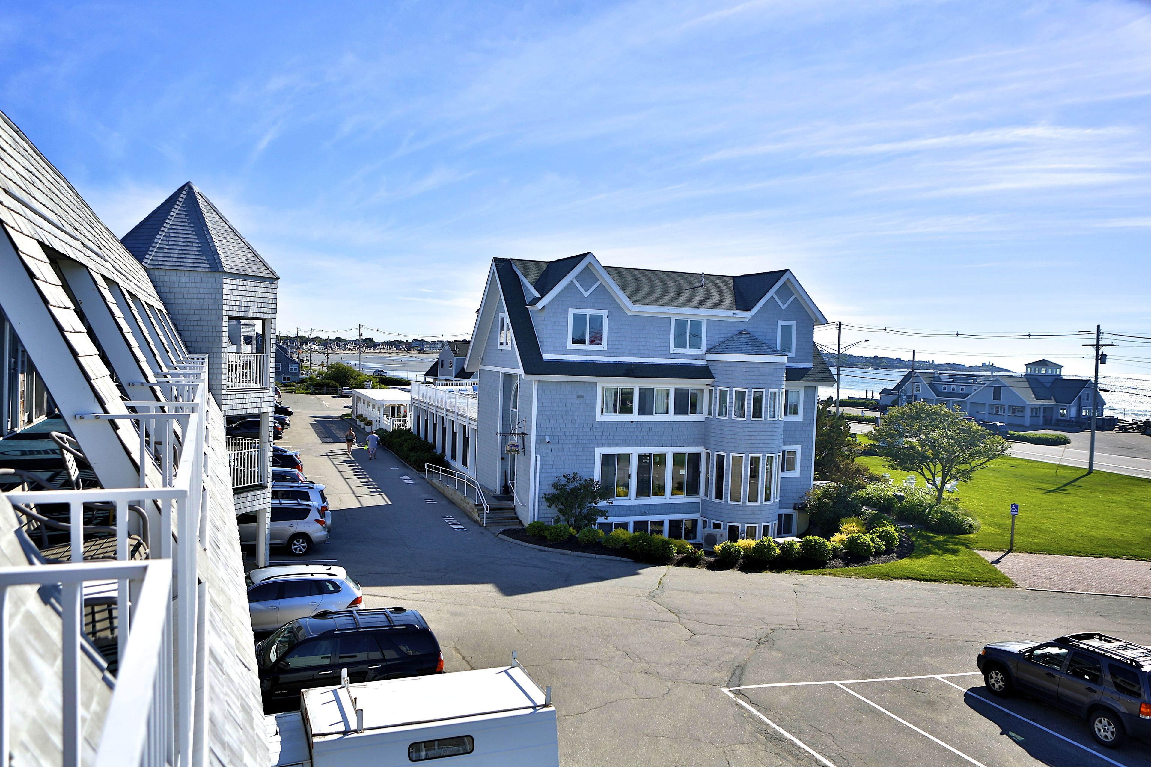 A large two-story building with a prominent gabled roof, a parking lot with several cars, and a clear blue sky.