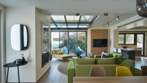 Uma sala de estar ampla com um sofá verde grande, um sofá amarelo e uma mesa de jantar. Há uma TV de tela plana na parede e um claraboia sobre a área de estar.