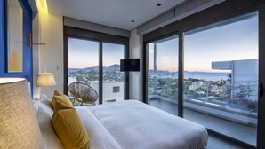 Quarto de hotel moderno com uma cama grande, televisão e vista da cidade através de janelas do chão ao teto.