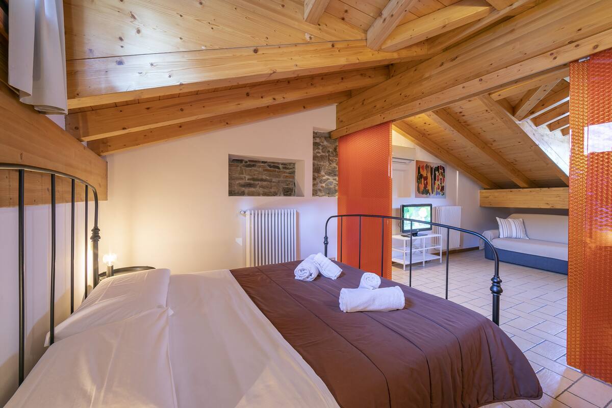 A bedroom with a wooden ceiling, a bed with white and brown bedding, a radiator, and a stone wall.