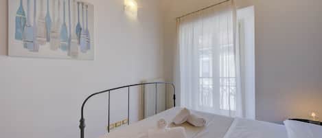 A neatly made bed with white linens and rolled towels, a metal headboard, a painting of bottles on the wall, and a window with sheer curtains.
