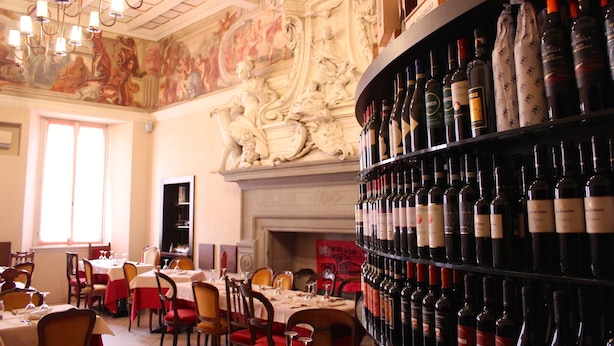 Un restaurante con elaborados adornos en el techo, una estantería con diversas botellas de vino y mesas dispuestas para comer.