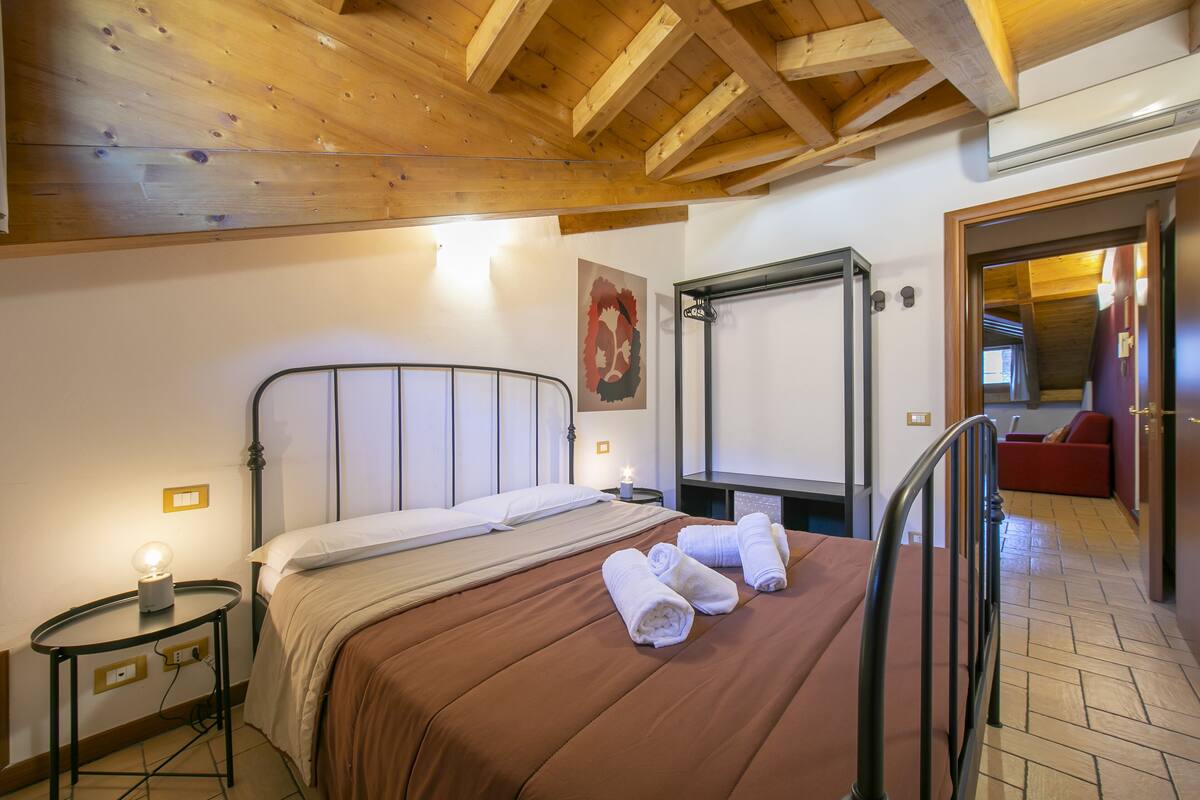 A bedroom with a wooden ceiling, a bed with a brown bedspread, two bedside tables with lamps, and a red sofa in the background.