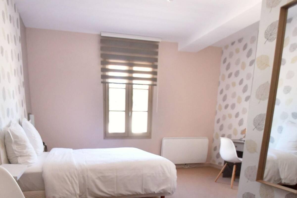 A bedroom with a bed, a desk, a chair, a window with blinds, and a mirror.