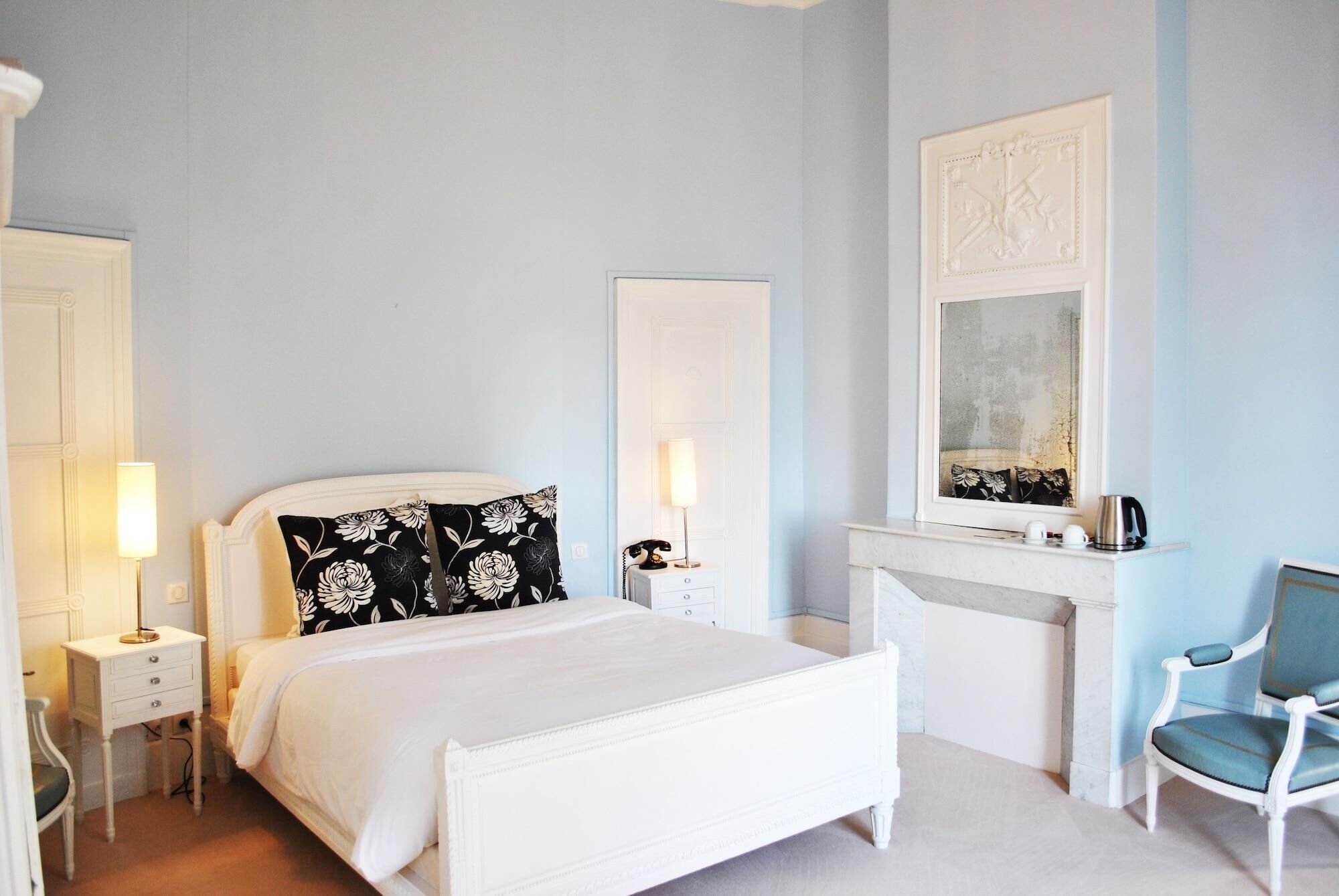 A bedroom with a white bed, bedside lamps, a chair, a fireplace, and a mirror.