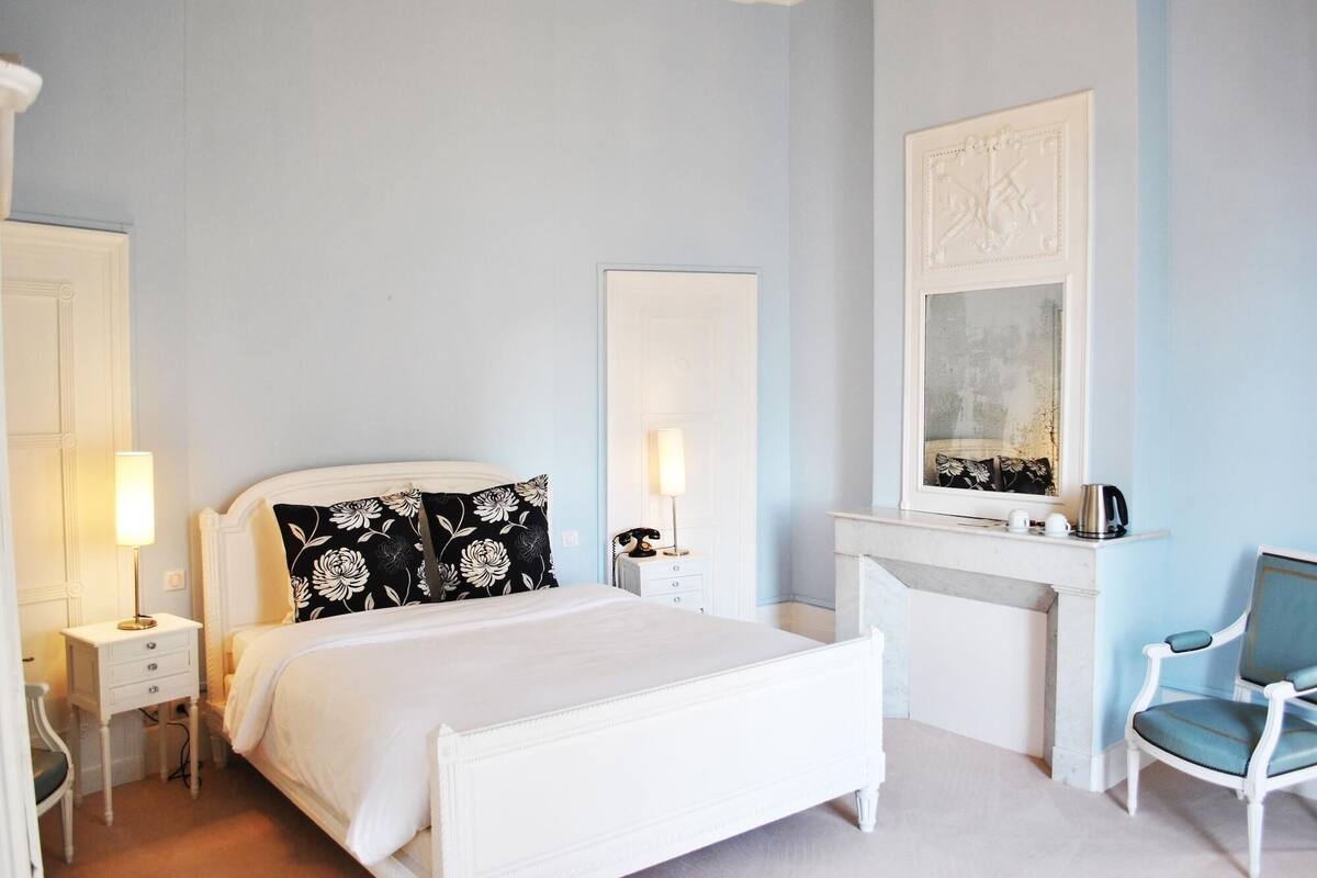 A bedroom with a white bed, bedside lamps, a chair, a fireplace, and a mirror.