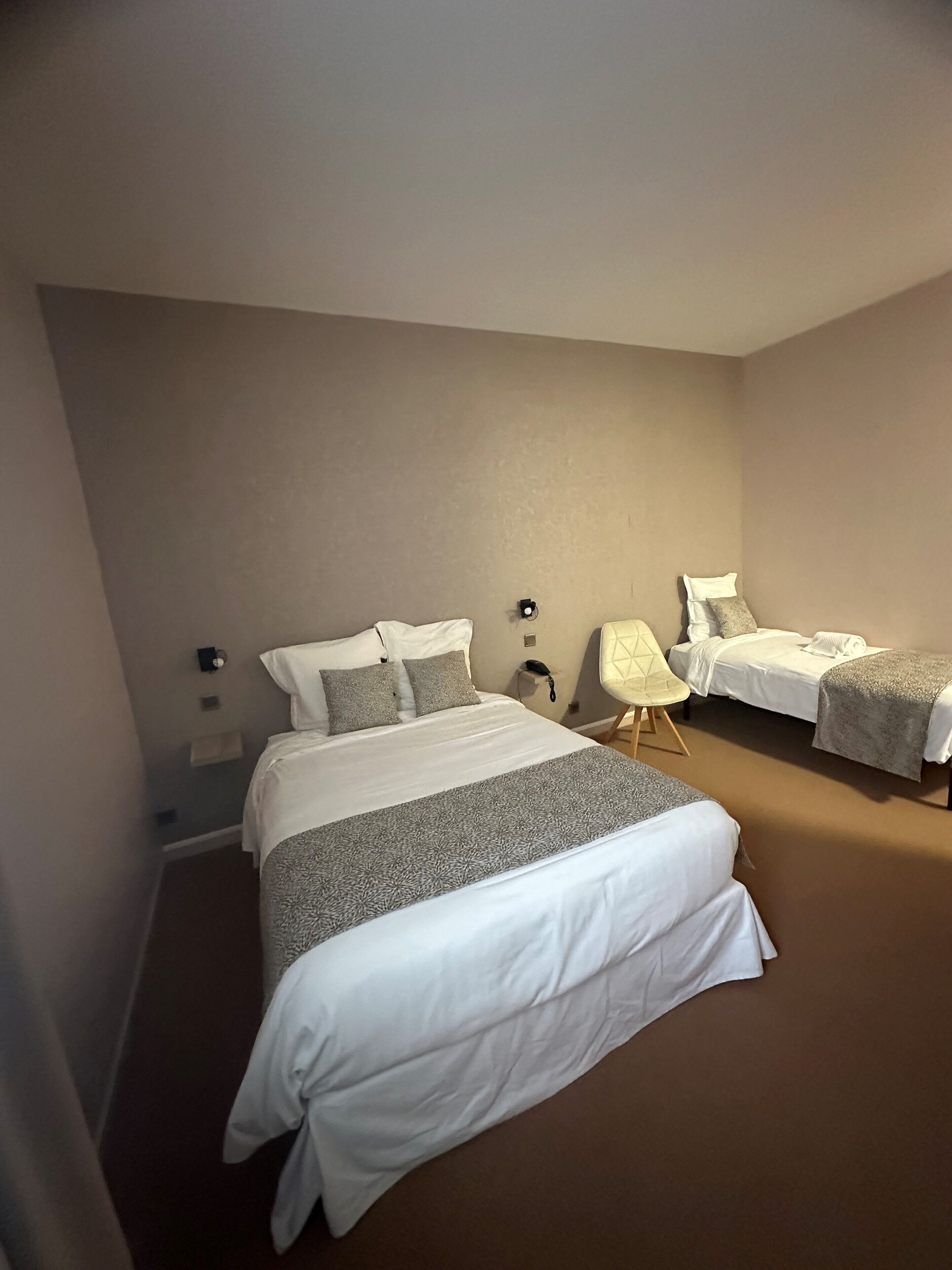A hotel room with two single beds, a bedside table with a lamp, a chair, and a wall-mounted phone.