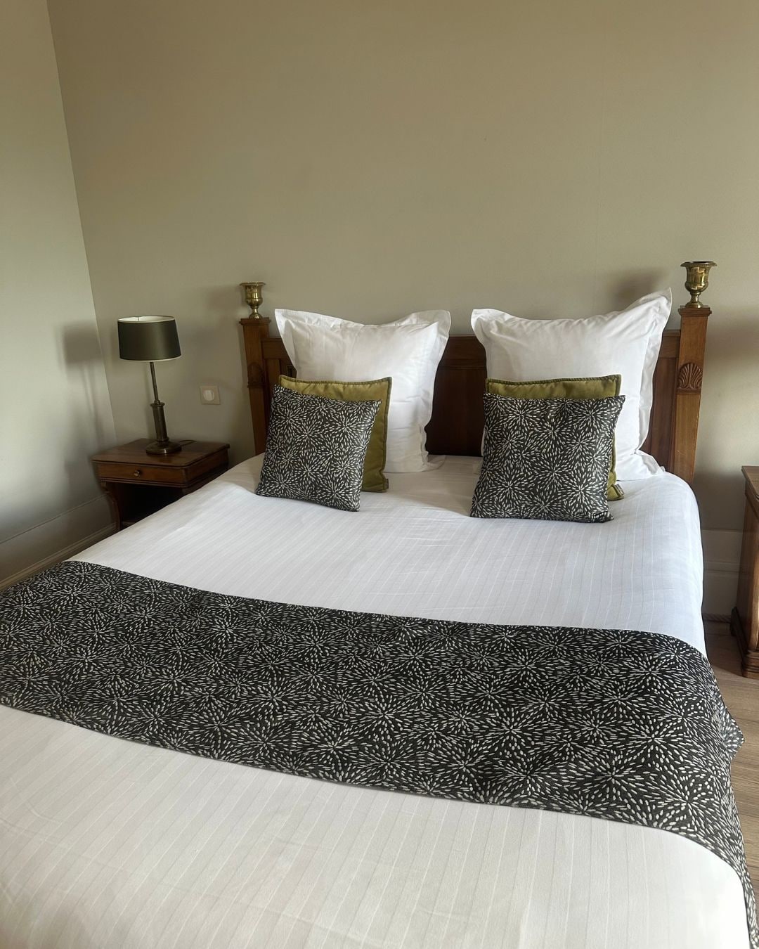 A neatly made bed with a white duvet and two decorative pillows, a bedside table with a lamp, and a wooden nightstand.