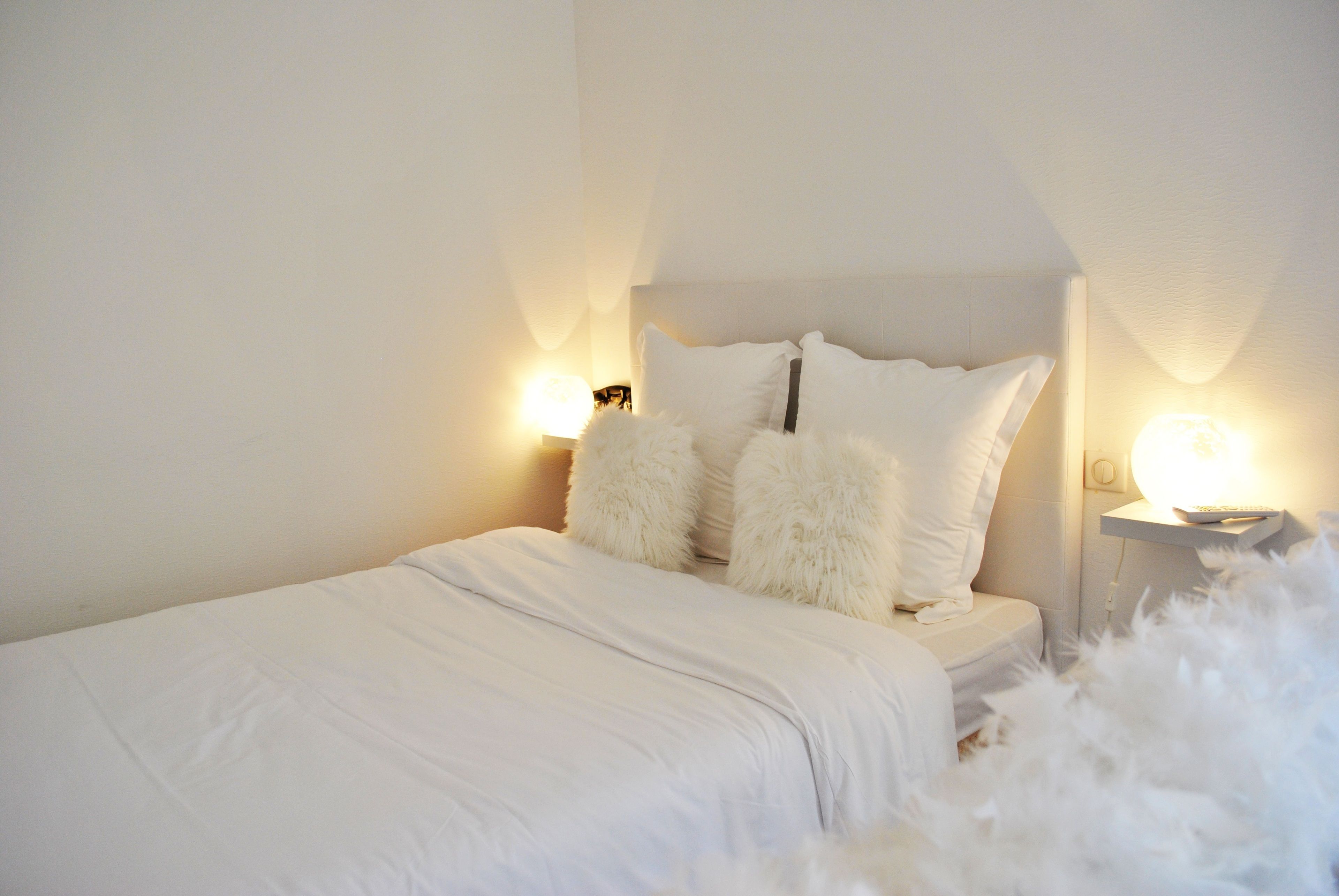 Una cama bien hecha con almohadas mullidas y un edredón blanco, dos lámparas de noche y un adorno mullido de color blanco en el suelo.