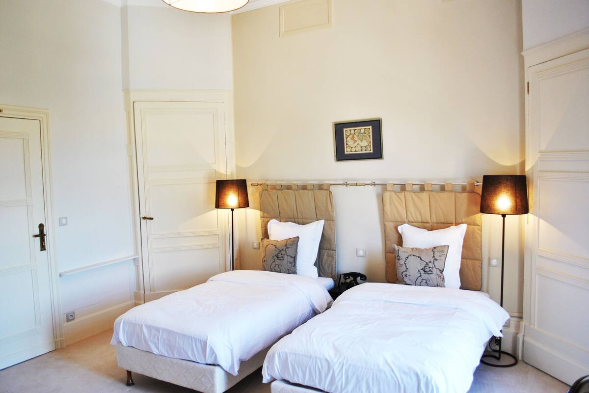A hotel room with two single beds, a headboard with a decorative panel, a framed artwork above the headboard, and two bedside lamps.