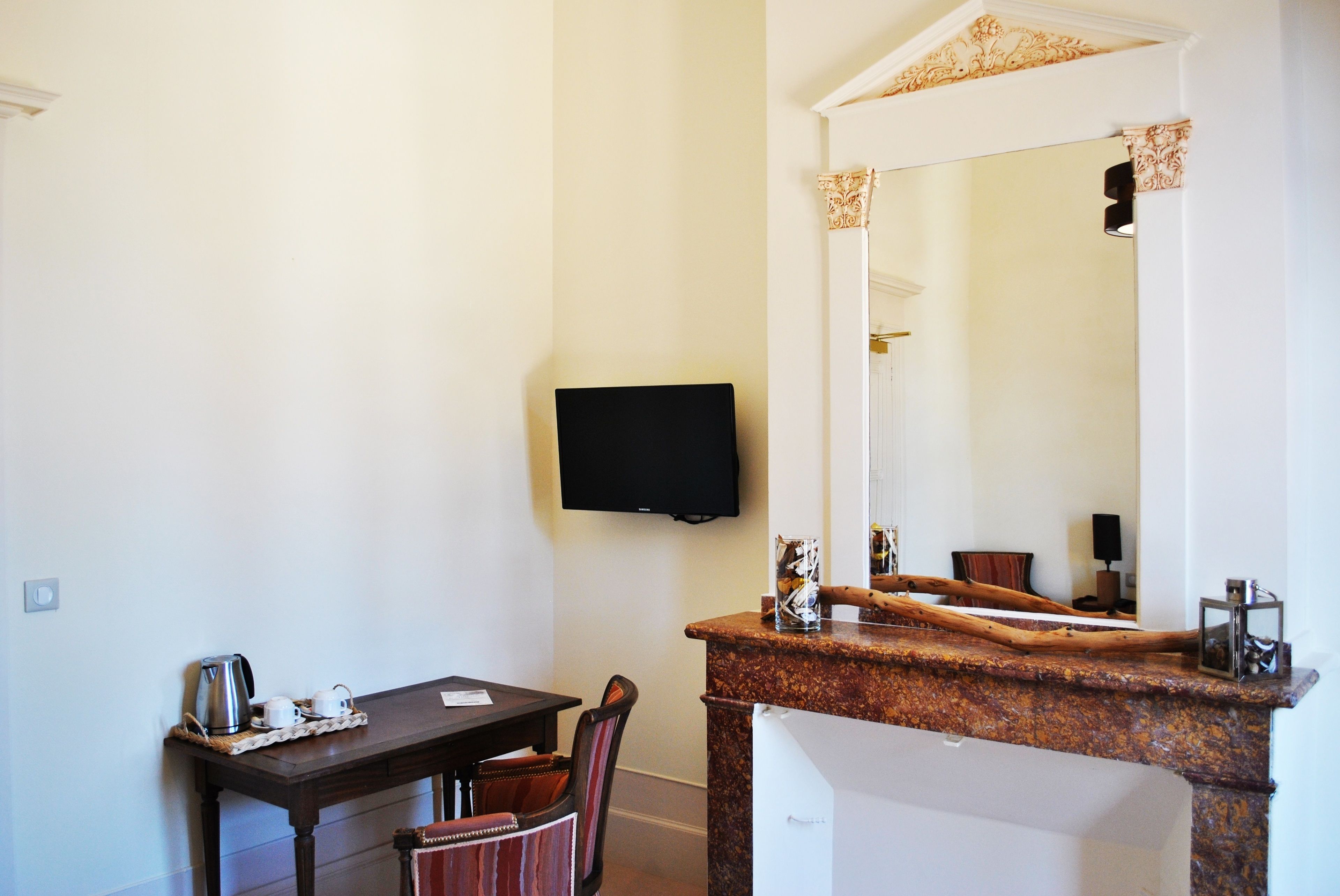 A hotel room with a wooden table, a chair, a television mounted on the wall, and a large mirror above a marble counter.